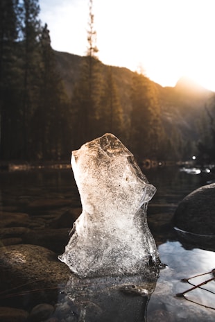 A block of ice stands upright in a shallow stream surrounded by smooth stones. The background features a forest with tall pine trees and a mountain silhouette under a brightly shining sun, casting warm light and long shadows.