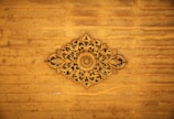 An intricate wooden carving with floral and leaf patterns centered against a distressed wooden panel background. The carving displays detailed craftsmanship with symmetrical designs radiating from a central floral motif.