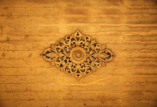 An intricate wooden carving with floral and leaf patterns centered against a distressed wooden panel background. The carving displays detailed craftsmanship with symmetrical designs radiating from a central floral motif.