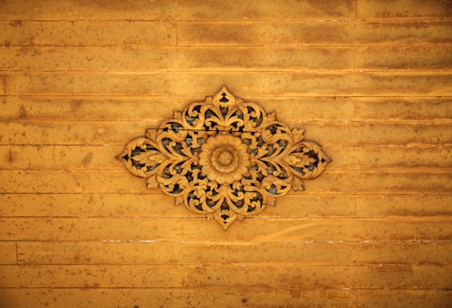 An intricate wooden carving with floral and leaf patterns centered against a distressed wooden panel background. The carving displays detailed craftsmanship with symmetrical designs radiating from a central floral motif.