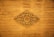 An intricate wooden carving with floral and leaf patterns centered against a distressed wooden panel background. The carving displays detailed craftsmanship with symmetrical designs radiating from a central floral motif.