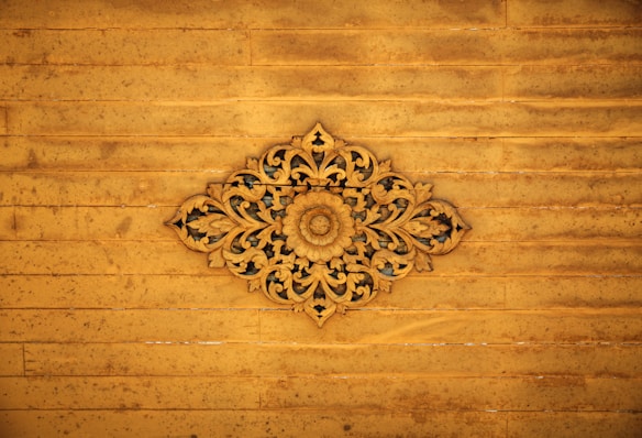An intricate wooden carving with floral and leaf patterns centered against a distressed wooden panel background. The carving displays detailed craftsmanship with symmetrical designs radiating from a central floral motif.
