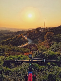 A picturesque landscape featuring a winding road through rolling hills. The sun sets on the horizon, casting a warm glow over the lush greenery. In the foreground, the handlebars and part of a mountain bike are visible, suggesting an outdoor adventure.