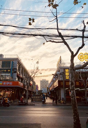 A busy city street with shopfronts on both sides selling various goods. Above, the sky is partly cloudy with a warm sunset, and several trees with sparse leaves line the street. Power lines crisscross the scene, adding an urban element. People are seen walking and riding scooters, suggesting a lively atmosphere. The lighting gives a warm, inviting feel to the image.