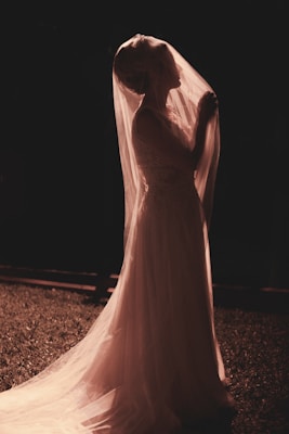 A bride in a delicate gown stands outdoors, bathed in soft, warm lighting. Her veil covers her face slightly, and she holds it gently with one hand as she gazes upwards. The background is dimly lit, adding to the serene and contemplative mood.