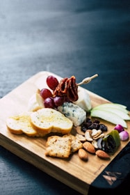 A wooden board displays a variety of snack items including sliced bread, green apple slices, grapes, various types of cheese, assorted nuts, olives, dried fruits, and crackers. A skewer with meat or vegetables is also visible, adding to the presentation.