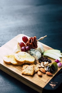 A wooden board displays a variety of snack items including sliced bread, green apple slices, grapes, various types of cheese, assorted nuts, olives, dried fruits, and crackers. A skewer with meat or vegetables is also visible, adding to the presentation.