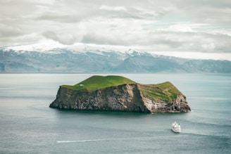 A green and rocky island sits in the middle of calm ocean waters, with a ferry or cruise ship sailing nearby. Snow-capped mountains are visible in the distance under a cloudy sky.