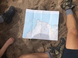 A map is spread out on sandy ground, surrounded by two people with hiking boots and one visible arm. The setting suggests planning or navigating during an outdoor adventure.