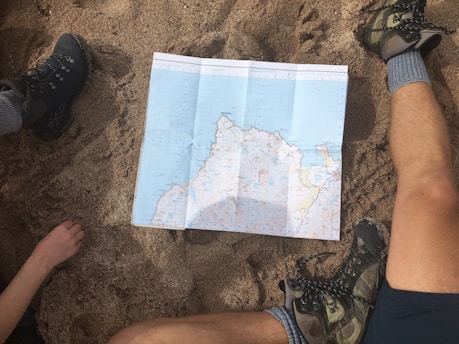 A map is spread out on sandy ground, surrounded by two people with hiking boots and one visible arm. The setting suggests planning or navigating during an outdoor adventure.