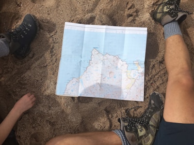 A map is spread out on sandy ground, surrounded by two people with hiking boots and one visible arm. The setting suggests planning or navigating during an outdoor adventure.