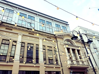 A classic building facade featuring large windows and decorative architectural details. String lights are draped across the area, adding a festive atmosphere. The building's soft beige and white color contrasts with the dark lines of the lights and fixtures.