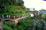 A group of people are hiking along a stone-paved trail surrounded by lush green vegetation. The path is elevated, leading up through the verdant landscape towards a small settlement of traditional buildings with blue and white accents. The hikers wear a variety of outdoor gear and backpacks.
