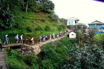 A group of people are hiking along a stone-paved trail surrounded by lush green vegetation. The path is elevated, leading up through the verdant landscape towards a small settlement of traditional buildings with blue and white accents. The hikers wear a variety of outdoor gear and backpacks.