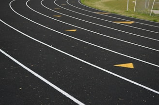 Several lanes of an outdoor running track are shown with curved white lines and yellow directional arrows on the surface. The track is bordered by green grass on one side, and a chain-link fence is visible in the background.