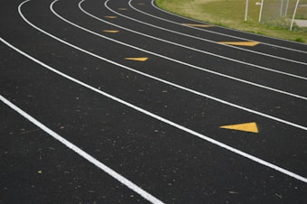 Several lanes of an outdoor running track are shown with curved white lines and yellow directional arrows on the surface. The track is bordered by green grass on one side, and a chain-link fence is visible in the background.