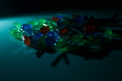 A collection of LED lights in various colors like green, blue, and orange are scattered on a surface. The scene is dimly lit, creating a soft glow and shadows, adding an atmospheric effect.