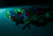A collection of LED lights in various colors like green, blue, and orange are scattered on a surface. The scene is dimly lit, creating a soft glow and shadows, adding an atmospheric effect.
