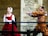 Two people dressed in historical clothing stand in front of a stone wall. One person, wearing a red dress and a white headscarf, has their hands clasped together. The other person, in a brown costume with metallic embellishments, holds a sword pointing it at the first person.