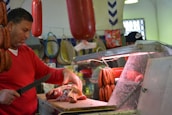 A butcher is working behind a meat counter, cutting a large piece of raw meat. Sausages and cured meat hang above the counter, and the man uses a knife to prepare the meat on a wooden board. The setting appears to be a market or a butcher shop with various meats and products in the background.