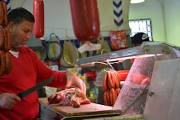 A butcher is working behind a meat counter, cutting a large piece of raw meat. Sausages and cured meat hang above the counter, and the man uses a knife to prepare the meat on a wooden board. The setting appears to be a market or a butcher shop with various meats and products in the background.