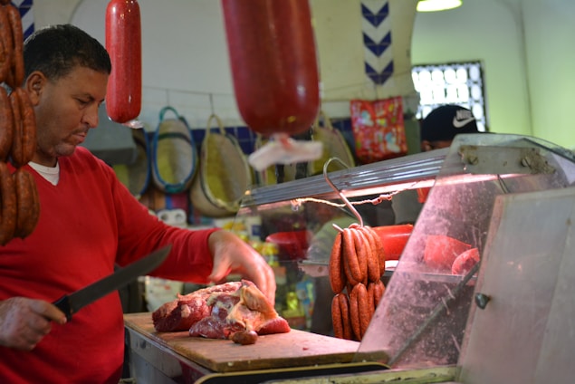 A butcher is working behind a meat counter, cutting a large piece of raw meat. Sausages and cured meat hang above the counter, and the man uses a knife to prepare the meat on a wooden board. The setting appears to be a market or a butcher shop with various meats and products in the background.
