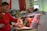 A butcher is working behind a meat counter, cutting a large piece of raw meat. Sausages and cured meat hang above the counter, and the man uses a knife to prepare the meat on a wooden board. The setting appears to be a market or a butcher shop with various meats and products in the background.