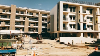 A building under construction with multiple floors and concrete columns. The area surrounding the building is an active construction site with equipment, materials, and workers visible. The façade of the structure features rectangular windows and balconies. The atmosphere appears dry and sunny.