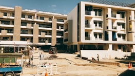 A building under construction with multiple floors and concrete columns. The area surrounding the building is an active construction site with equipment, materials, and workers visible. The façade of the structure features rectangular windows and balconies. The atmosphere appears dry and sunny.