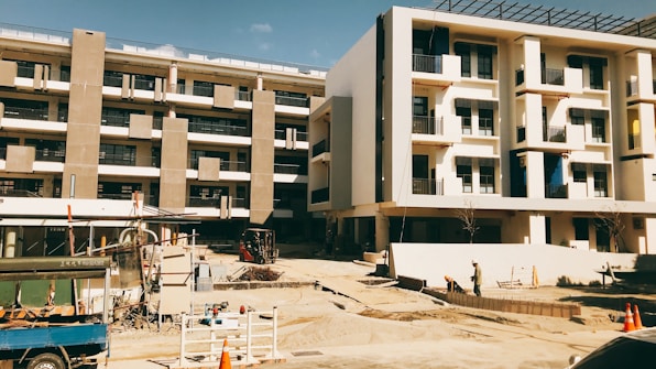 A building under construction with multiple floors and concrete columns. The area surrounding the building is an active construction site with equipment, materials, and workers visible. The façade of the structure features rectangular windows and balconies. The atmosphere appears dry and sunny.