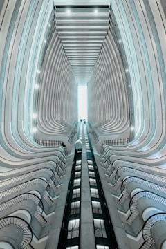 A modern architectural structure with an upward perspective showing multiple floors. The view encompasses two elevators in motion towards a bright skylight. Curved balconies line the sides, creating a sense of depth and symmetry.