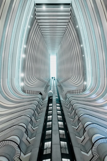 A modern architectural structure with an upward perspective showing multiple floors. The view encompasses two elevators in motion towards a bright skylight. Curved balconies line the sides, creating a sense of depth and symmetry.