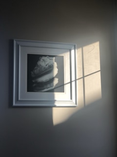 A framed black and white photograph hangs on a wall, partially illuminated by sunlight streaming through a window. The frame is white and the sunlight creates dynamic shadows across both the wall and the photograph.