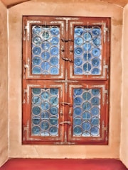 A window with intricate metalwork and decorative blue glass arranged in circular patterns. The window is framed with ornate wooden borders, complemented by ornate metal hinges and handles. The surrounding wall has a smooth, beige finish.