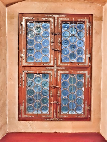 A window with intricate metalwork and decorative blue glass arranged in circular patterns. The window is framed with ornate wooden borders, complemented by ornate metal hinges and handles. The surrounding wall has a smooth, beige finish.