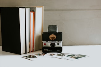 A vintage instant camera is placed on a surface next to a stack of books. Several instant photographs are scattered in front of the camera, adding a nostalgic feel to the scene. The background features a textured, light-colored wall that complements the overall aesthetic.