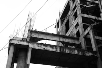 An incomplete concrete building structure with exposed rebars and several partially constructed floors. The photograph is in black and white, highlighting the stark, unfinished nature of the building. Several cables or wires are visible, intersecting the image diagonally.