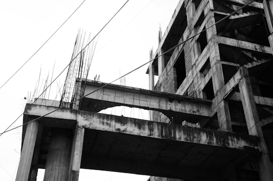 An incomplete concrete building structure with exposed rebars and several partially constructed floors. The photograph is in black and white, highlighting the stark, unfinished nature of the building. Several cables or wires are visible, intersecting the image diagonally.