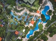 An aerial view of a large, curvaceous swimming pool complex surrounded by lush greenery and landscaped gardens. The pools are bordered by stone pathways and flanked by verdant trees and neatly arranged plants. Several small structures with red roofs can be seen, along with sun loungers and umbrellas spread around the pool areas.