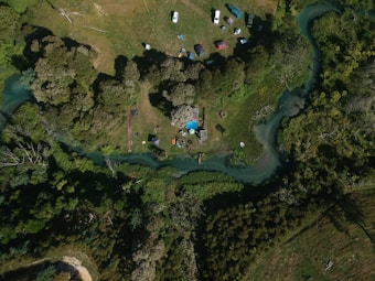 An aerial view of a lush, green forest area with a winding river that curves through the landscape. The image includes several vehicles and tents scattered around a small, open clearing. There are tall trees casting shadows over parts of the grassy field.
