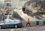 A mural resembling a classic painting is depicted on the side of a building, incorporating elements like a car parked in front. A person wearing an orange safety jacket and a hard hat is walking past, and there are trash bins nearby. The mural is inspired by a famous artwork and features dynamic movement of figures reaching out towards each other.