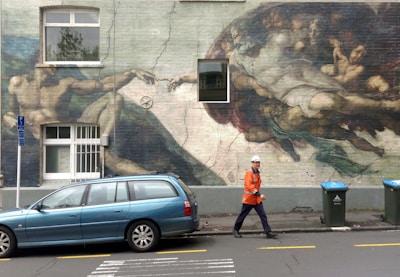 A mural resembling a classic painting is depicted on the side of a building, incorporating elements like a car parked in front. A person wearing an orange safety jacket and a hard hat is walking past, and there are trash bins nearby. The mural is inspired by a famous artwork and features dynamic movement of figures reaching out towards each other.