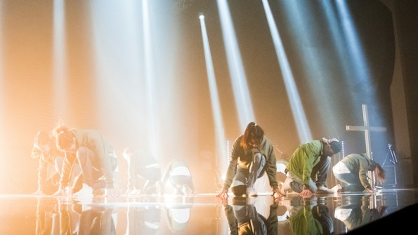 Several individuals are kneeling on a stage with dramatic lighting, creating shadows and reflections on the floor. Beams of light create a dynamic effect, suggesting a performance or rehearsal setting. The atmosphere is intensified by the presence of a large cross and musical equipment in the background.