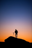 silhouette of person standing beside DSLR camera with stand at sunset