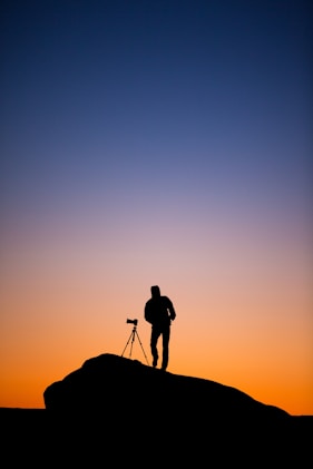 silhouette of person standing beside DSLR camera with stand at sunset