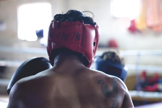 A person wearing a red protective headgear is shown from the back in a boxing or training environment. The setting appears to be a gym, with blurred boxing rings visible in the background. On the person's shoulder, there is a visible tattoo of a rooster.