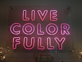 Bright pink neon sign with the words 'LIVE COLOR FULLY' mounted on a transparent panel. The sign is illuminated in a dimly lit setting with decorative lights partially visible on the sides.