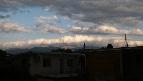 A residential rooftop scene with a view of distant mountains under a partially cloudy sky. The clouds are thick and fluffy near the horizon, with patches of blue sky visible. The structures in the foreground appear to be houses with flat rooftops, and there are a few plant shrubs visible.