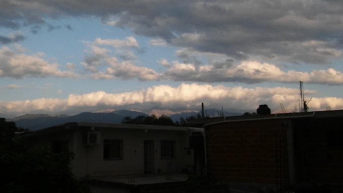 A residential rooftop scene with a view of distant mountains under a partially cloudy sky. The clouds are thick and fluffy near the horizon, with patches of blue sky visible. The structures in the foreground appear to be houses with flat rooftops, and there are a few plant shrubs visible.