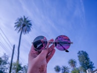 A hand holding a pair of round sunglasses with reflection showing various scenes, against a blue sky with palm trees. The sunglasses lenses have a purple tint that contrasts with the clear sky and greenery.